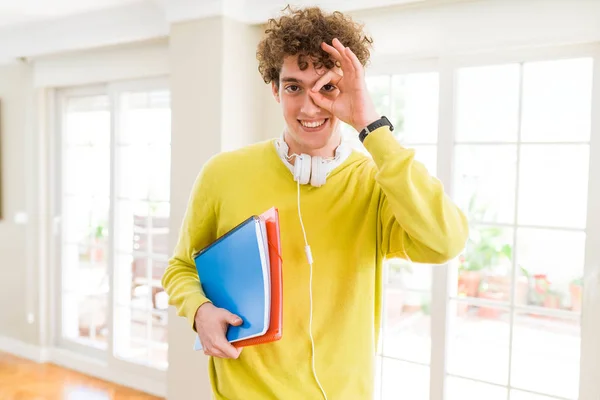 年轻的学生男子戴着耳机 拿着笔记本 开心的脸微笑着做好标志 手对眼睛透过手指看 — 图库照片