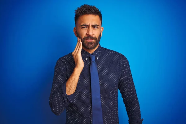 Young indian businessman wearing elegant shirt and tie standing over isolated blue background touching mouth with hand with painful expression because of toothache or dental illness on teeth. Dentist concept.