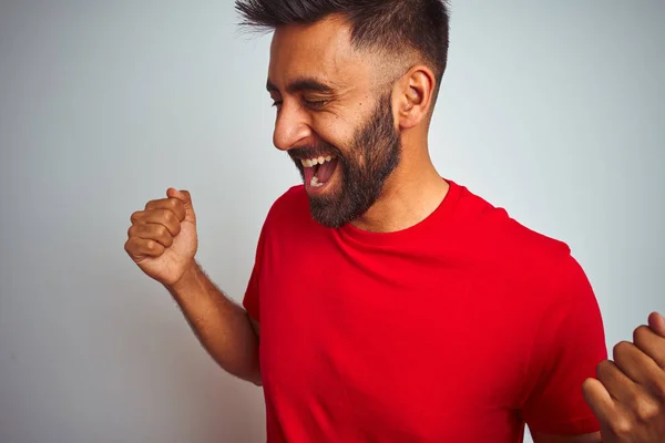 Young indian man wearing red t-shirt over isolated white background very happy and excited doing winner gesture with arms raised, smiling and screaming for success. Celebration concept.