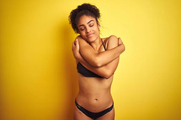 African american woman on vacation wearing bikini standing over isolated yellow background Hugging oneself happy and positive, smiling confident. Self love and self care