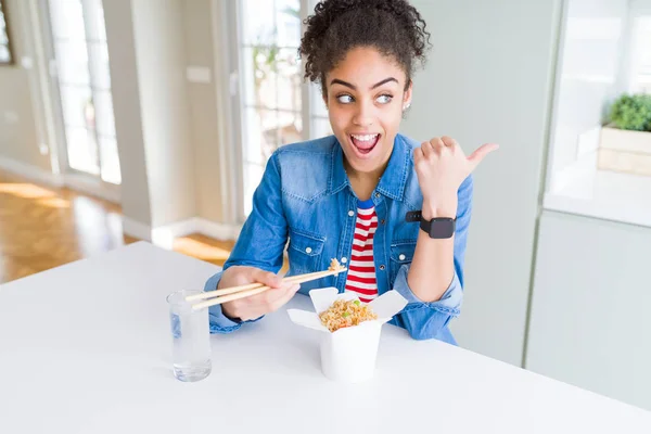 Young african american woman eating asian noodles from delivery box pointing and showing with thumb up to the side with happy face smiling