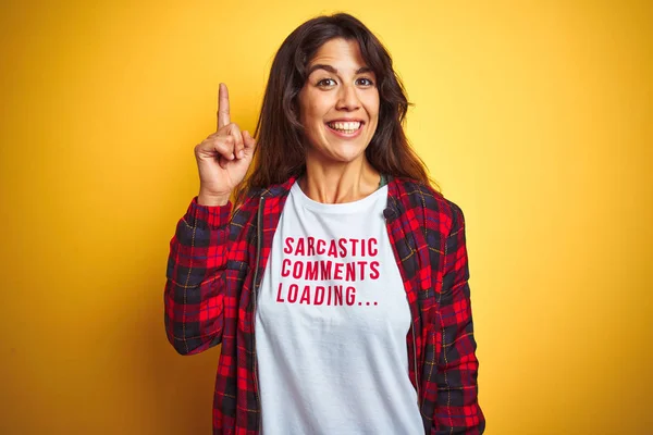 Beautiful woman wearing funny t-shirt with irony comments over isolated yellow background surprised with an idea or question pointing finger with happy face, number one