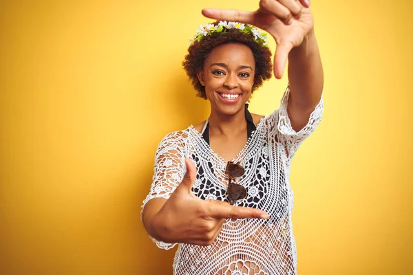 Young african american woman with afro hair wearing flowers crown over yellow isolated background smiling making frame with hands and fingers with happy face. Creativity and photography concept.