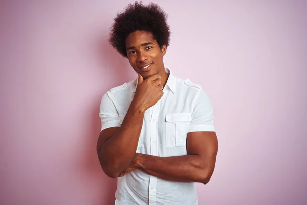 Young american man with afro hair wearing white shirt standing over isolated pink background looking confident at the camera with smile with crossed arms and hand raised on chin. Thinking positive.