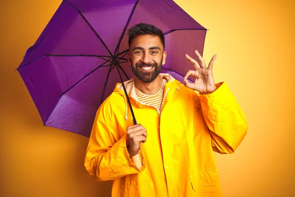 Young indian man wearing raincoat and purple umbrella over isolated yellow background doing ok sign with fingers, excellent symbol