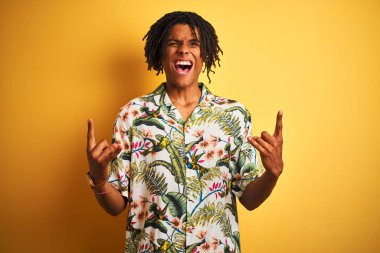 Tatil üzerinde dreadlocks ile Afro adam izole sarı arka plan üzerinde el ile kaya sembolü yapıyor çılgın ifade bağırarak üzerinde yaz gömlek giyiyor. Müzik yıldızı. Ağır kavram.