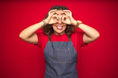 Orta yaş kıdemli kadın kırmızı izole arka plan üzerinde önlük üniforma giyiyor dürbün yapışması gibi ok jest yapıyor, gözler parmaklarla bakıyor. Çılgın ifade.