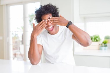Genç afro-amerikan adam rahat beyaz t-shirt giyen evde elleri neşeli ve komik gülümseyerek gözleri kaplayan oturuyor. Kör kavramı.