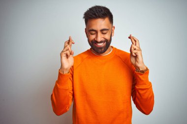 İzole beyaz arka plan üzerinde turuncu kazak giyen genç Hintli adam parmak umut ve gözler ile gülümseyen geçti kapalı. Şans ve batıl fikir.