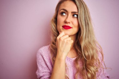 Pembe izole arka plan üzerinde ayakta kazak giyen genç güzel kadın soru hakkında düşünme ciddi yüz, çok karışık fikir