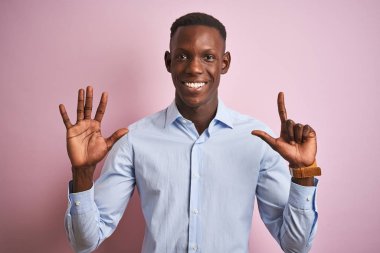 İzole pembe arka plan üzerinde ayakta mavi zarif gömlek giyen Afrikalı Amerikalı adam gösteren ve parmak sayısı yedi ile işaret kadar güven ve mutlu gülümseyen.