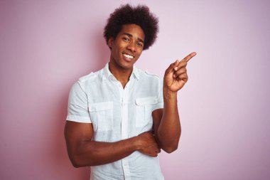 Afro saç ile genç Amerikan adam yüz üzerinde büyük bir gülümseme ile izole pembe arka plan üzerinde ayakta beyaz gömlek giyen, kamera bakarak yan el ve parmak ile işaret.