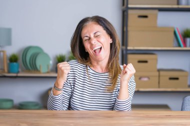 Orta yaş kıdemli kadın evde masada oturan çok mutlu ve heyecanlı silah ile kazanan jest yaparak, gülümseyen ve başarı için çığlık kaldırdı. Kutlama konsepti.