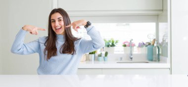 Evde beyaz masada oturan güzel genç kadının geniş açı resim, parmaklar ve ağız ile gösteren ve işaret emin gülümseyen. Sağlık konsepti.