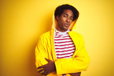 Afro saç, izole sarı arka plan üzerinde Hood ayakta kaput ile yağmur ceket giyen Afrikalı adam kamera bakarak çapraz silah ile gülümseyen yüz. Pozitif kişi.