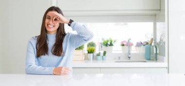 Geniş açı resim güzel genç kadının evde beyaz masaya oturan gülümsüyor, parmakları ile mutlu yüz üzerinden seyir göz el ile ok jest yapıyor.