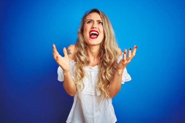 Beyaz elbise giyen genç güzel kadın mavi izole arka plan deli ve deli bağırma ve agresif ifade ve silah ile bağırarak kaldırdı. Hayal kırıklığı kavramı.