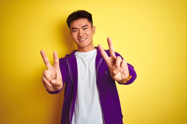 Genç Asyalı Çinli adam izole sarı arka plan üzerinde duran mor Sweatshirt ayakta giyen kamera parmaklar zafer işareti yapıyor gösteren bakıyor. İki numara.