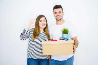 Beyaz izole arka plan üzerinde yeni bir ev taşımak karton kutu tutan genç çift parmaklar ile ok işareti, mükemmel sembolü yapıyor