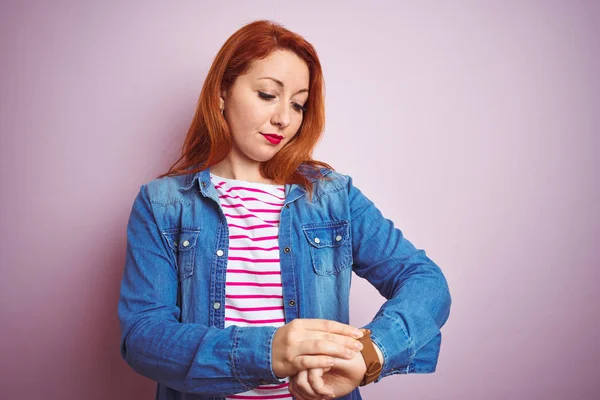 Beautiful redhead woman wearing denim shirt and striped t-shirt over isolated pink background Checking the time on wrist watch, relaxed and confident