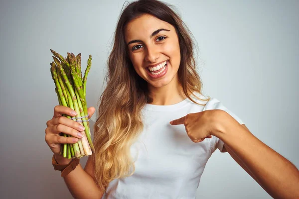 灰色の孤立した背景の上にアスパラガスを食べる若い美しい女性は 驚きの顔が自分自身に指を指さす — ストック写真