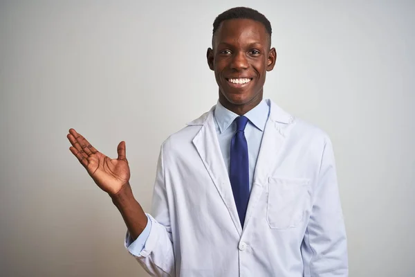 Young african american doctor man wearing coat standing over isolated white background smiling cheerful presenting and pointing with palm of hand looking at the camera.