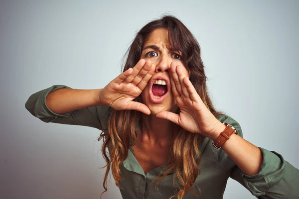 Young beautiful woman wearing green shirt standing over grey isolated background Shouting angry out loud with hands over mouth