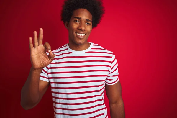 孤立した赤い背景の上にストライプのTシャツを着たアフロヘアの若いアフリカ系アメリカ人男性は 手と指でOkサインをやって肯定的に微笑んでいます 成功した式 — ストック写真