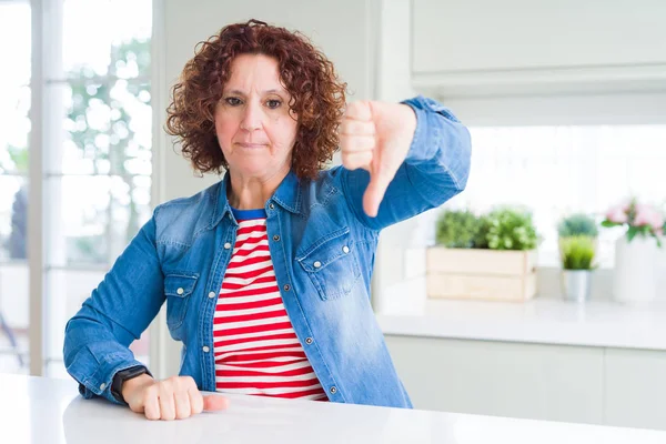Evde kot ceket giyen kıvırcık saçlı orta yaş kıdemli kadın mutsuz ve öfkeli gösteren reddetme ve başparmak aşağı jest ile negatif görünümlü. Kötü ifade.