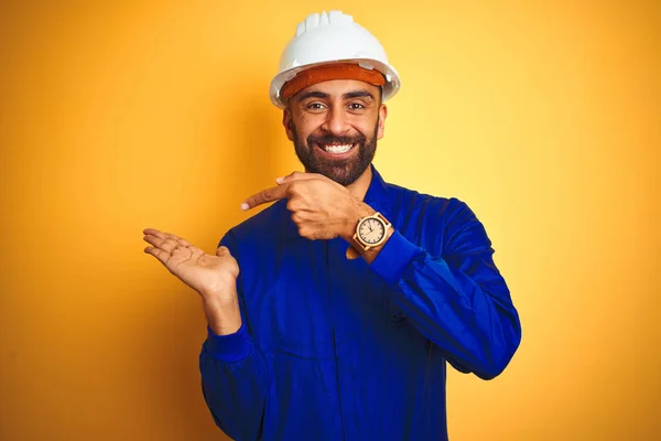 Handsome indian worker man wearing uniform and helmet over isolated yellow background amazed and smiling to the camera while presenting with hand and pointing with finger.