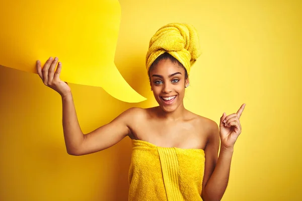 Afro woman wearing towel after shower holding speech bubble over isolated yellow background very happy pointing with hand and finger to the side