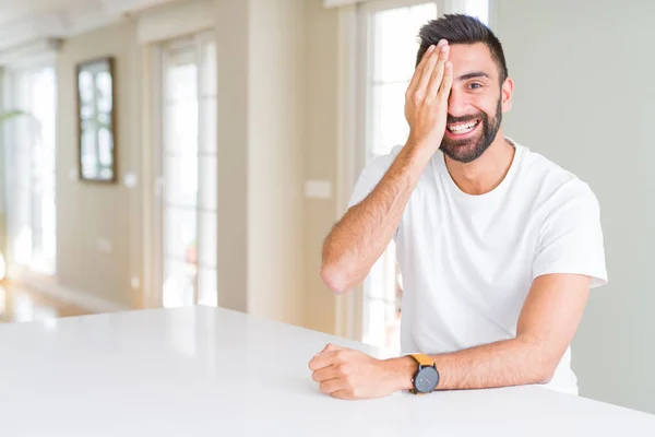英俊的西班牙男子休闲白色T恤在家里覆盖一只眼睛与自信的笑容在脸上和惊喜的情绪 — 图库照片