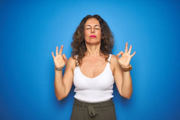 青い孤立した背景の上に立つ巻き毛を持つ中年のシニア女性は 指で瞑想ジェスチャーをして目を閉じて微笑んでいます ヨガのコンセプト — ストック写真