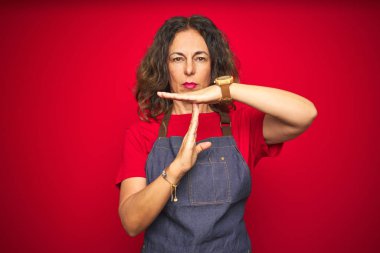 Kırmızı izole arka plan üzerinde apron üniforma giyen orta yaş kıdemli kadın eller, sinirli ve ciddi yüz ile zaman aşımına jest yapıyor