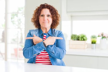 Evde kot ceket giyen kıvırcık saçlı orta yaş kıdemli kadın parmaklarıyla her iki tarafa Işaret, farklı yönde katılmıyorum