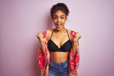 Tatil üzerinde bikini giyen Amerikan kadın ve izole pembe arka plan üzerinde renkli elbise şaşırttı ve silah kaldırdı ve açık gözleri ile başarı için hayran kutluyor. Kazanan kavramı.