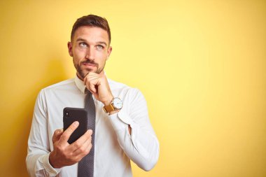 Sarı izole arka plan üzerinde akıllı telefon kullanarak genç yakışıklı iş adamı soru hakkında düşünme ciddi yüz, çok karışık fikir