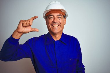 Yakışıklı orta yaş Worker adam üniforma ve kask üzerinde izole beyaz arka plan gülümseyen ve el ile küçük boyutu işareti yapıyor parmak görünümlü ve kamera ile emin işaret giyiyor. Ölçü kavramı.