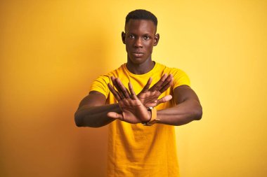 İzole sarı arka plan üzerinde ayakta rahat t-shirt giyen genç Afrikalı Amerikalı adam ret ifadesi geçiş silah ve avuç içi negatif işareti, öfkeli yüz yapıyor