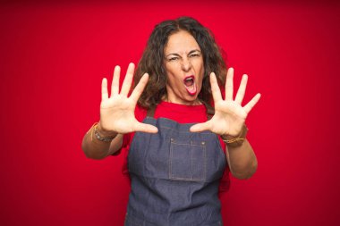 Orta yaş kıdemli kadın el avuç içi, öfkeli ve hayal kırıklığı ifade ile stop jest yapıyor kırmızı izole arka plan üzerinde apron üniforma giyiyor