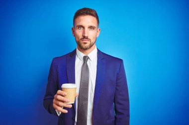 Genç yakışıklı iş adam ciddi düşünme akıllı yüz üzerinde güvenli bir ifade ile mavi izole arka plan üzerinde bir kağıt fincan bir kahve içme