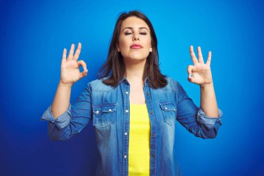 Mavi izole arka plan üzerinde Casual mavi denim ceket giyen genç güzel esmer kadın Relax ve gözleri ile gülümseyen meditasyon jest parmaklar ile kapalı. Yoga konsepti.