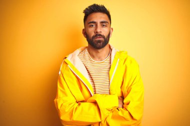 Genç Hint adam izole sarı arka plan üzerinde duran yağmurluk giyen şüpheci ve sinir, kesişmiş kollar ile yüz üzerinde ifade onaylamıyor. Negatif kişi.