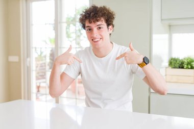 Evde beyaz tişört giyen genç yakışıklı adam yüzünde gülümseme ile kendinden emin görünümlü, parmakları gururlu ve mutlu kendini işaret.