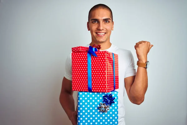 Young handsome man holding birthday gifts over white isolated background pointing and showing with thumb up to the side with happy face smiling