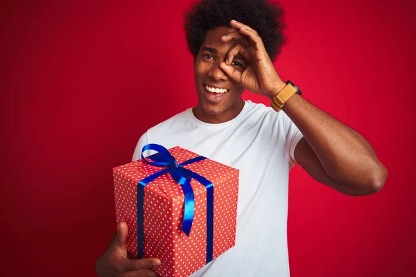 Young african american man holding birthday gift standing over isolated red background with happy face smiling doing ok sign with hand on eye looking through fingers