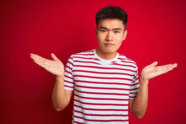 孤立した赤い背景の上に立つストライプのTシャツを着た若いアジア人の中国人男性は 腕と手を上げて混乱した表情を浮かべた 疑いの概念 — ストック写真