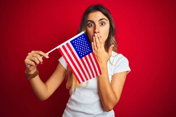 Young woman holding united states of america flag over red isolated background cover mouth with hand shocked with shame for mistake, expression of fear, scared in silence, secret concept
