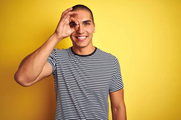 年轻的帅哥穿着条纹T恤在黄色孤立的背景做Ok手势与手微笑 眼睛透过手指看与快乐的脸 — 图库照片