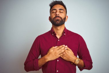 Kırmızı zarif gömlek giyen genç Hint adam izole gri arka planda ayakta gözleri ve yüz minnettar jest ile göğsüne eller ile gülümseyen. Sağlık konsepti.